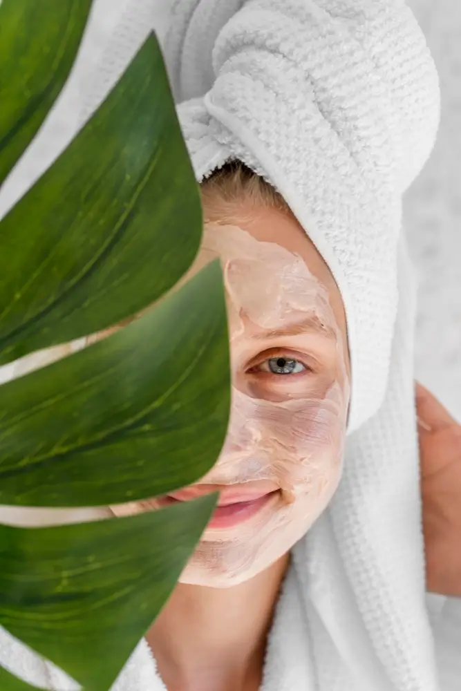 Femme souriante avec un masque de soin sur le visage partiellement caché par une grande feuille verte et une serviette de bain sur la tête